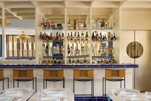 A bar area with multiple shelves of bottles, bar stools, and tables set for dining. - Fenix Hotel (Glyfada)