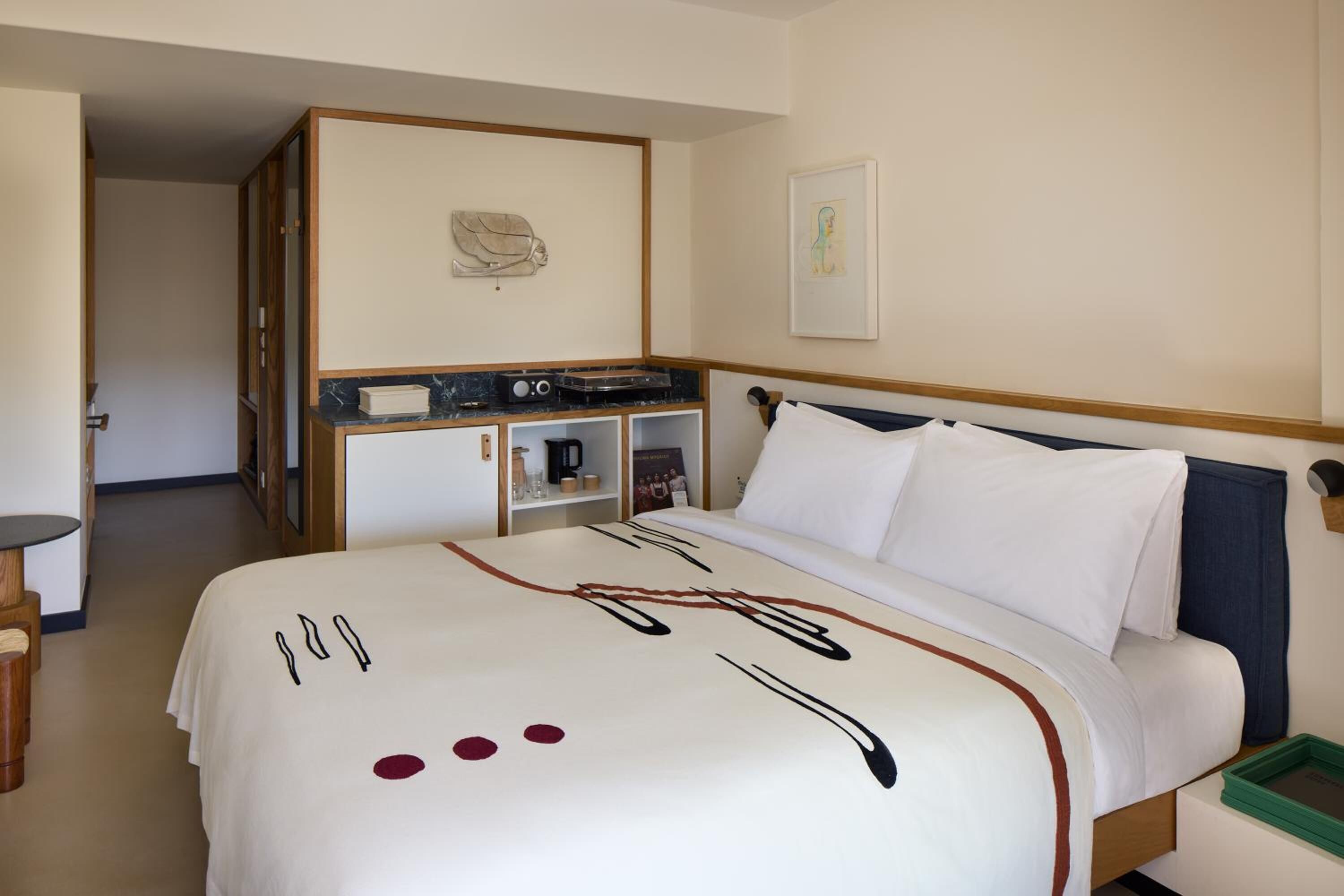 A hotel room with a bed, a desk, a chair, a coffee machine, and a framed picture on the wall.