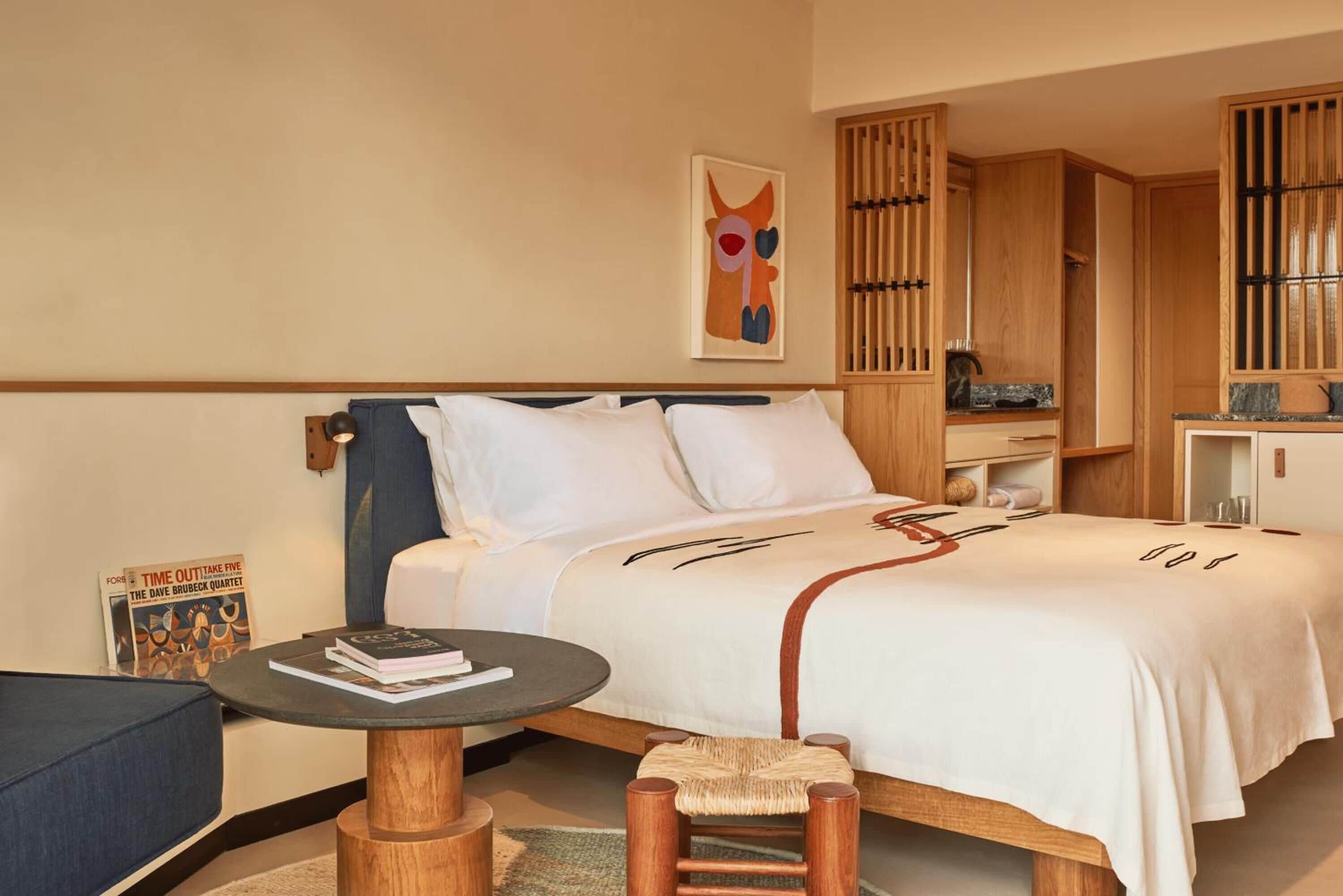 A hotel room with a bed, a small round table, a chair, a bookshelf, and a painting on the wall.