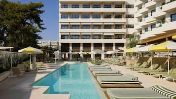 A swimming pool with lounge chairs and umbrellas, a multi-story building with balconies, and a clear sky.