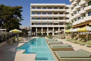 A swimming pool with lounge chairs and umbrellas, a multi-story building with balconies, and a clear sky. - Fenix Hotel (Glyfada)