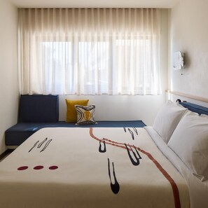 A hotel room with a bed, a window with sheer curtains, a sofa with cushions, and a wall-mounted light fixture. - Fenix Hotel (Glyfada)