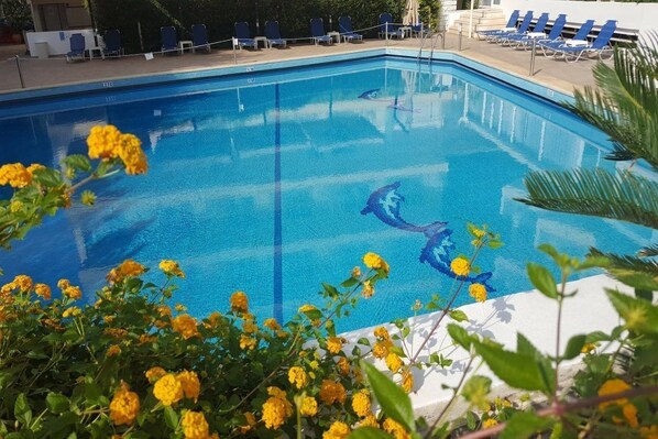 A swimming pool with clear blue water, surrounded by lounge chairs and yellow flowers. - Fenix Hotel (Glyfada)