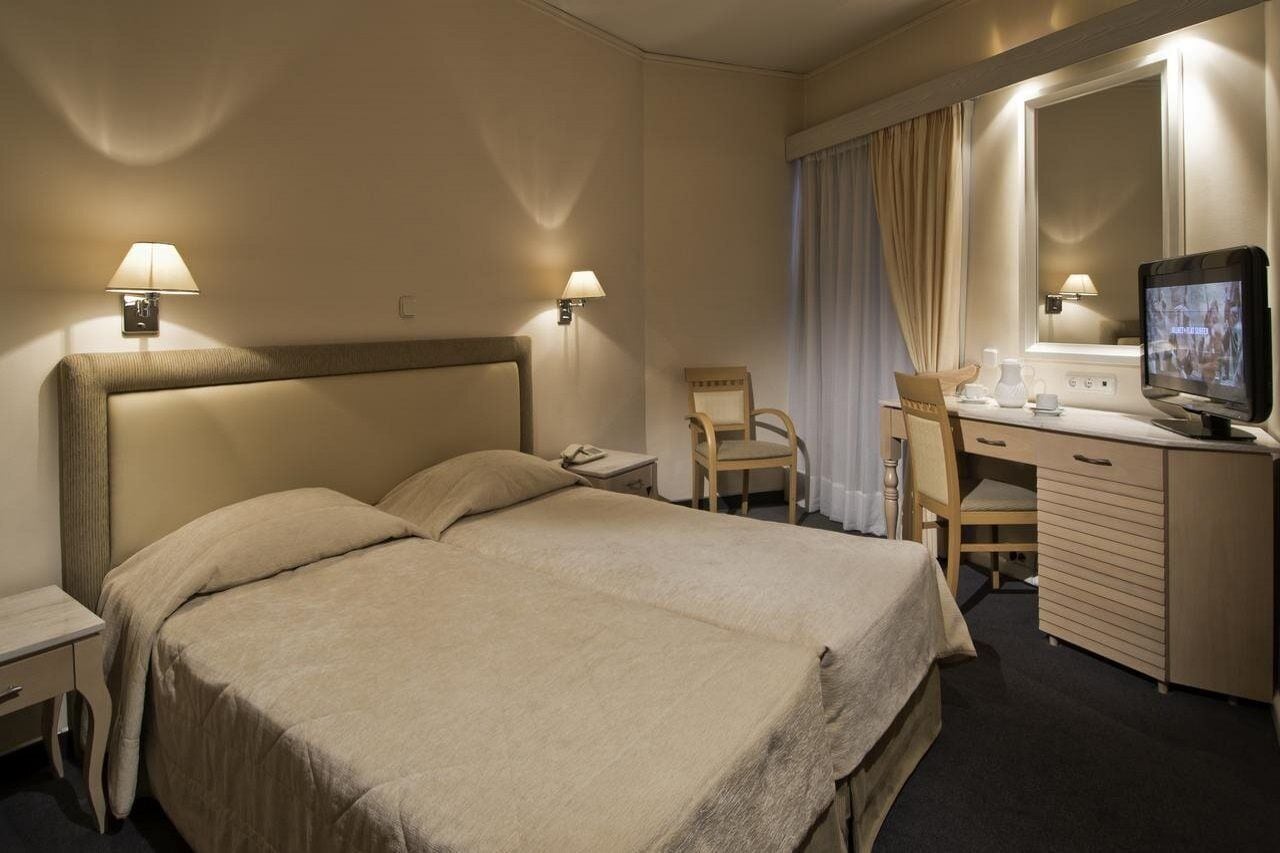 A hotel room with a bed, a desk, a chair, a TV, and two wall-mounted lamps.