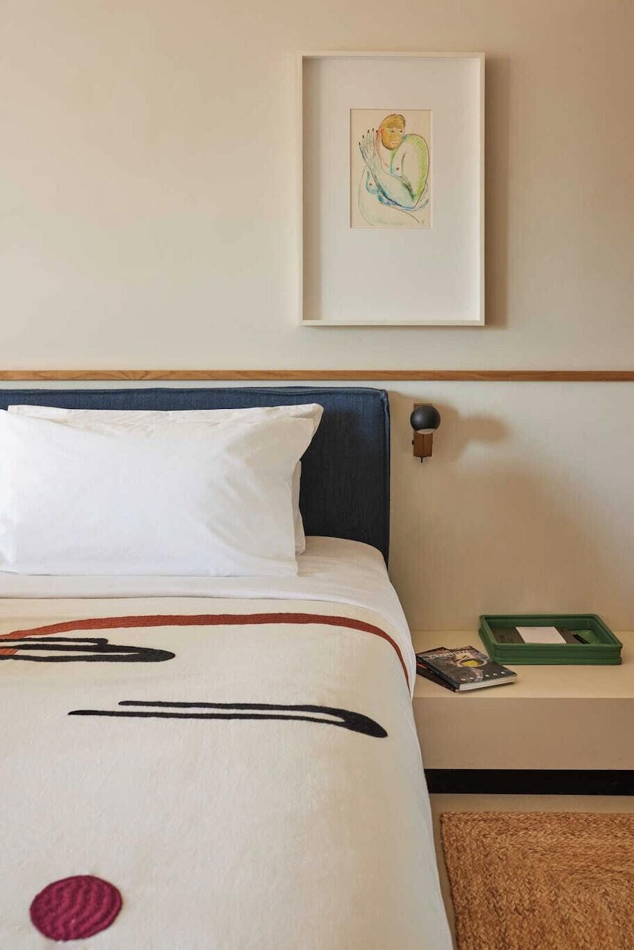 Et hotelværelse med en seng, et billede hængende på væggen, et natbord med en bog og en grøn bakke, samt et brunt tæppe.