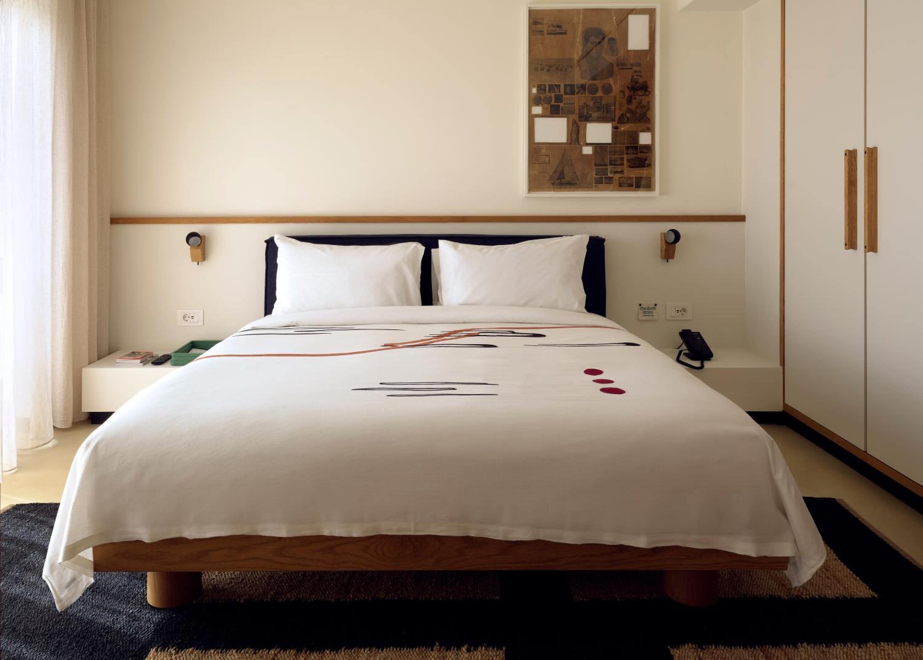 A neatly made bed with a white bedspread, a bedside table with a book and remote, and a wall-mounted artwork above the bed.
