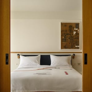 Hotellihuone, jossa on sänky, sängynpääty, seinälle ripustettu taideteos ja puuovi.