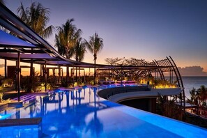 Área de piscina com borda curva, palmeiras e vista para o oceano ao entardecer.