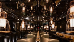 A modern interior with a central long table, multiple seating areas, and unique hanging light fixtures.