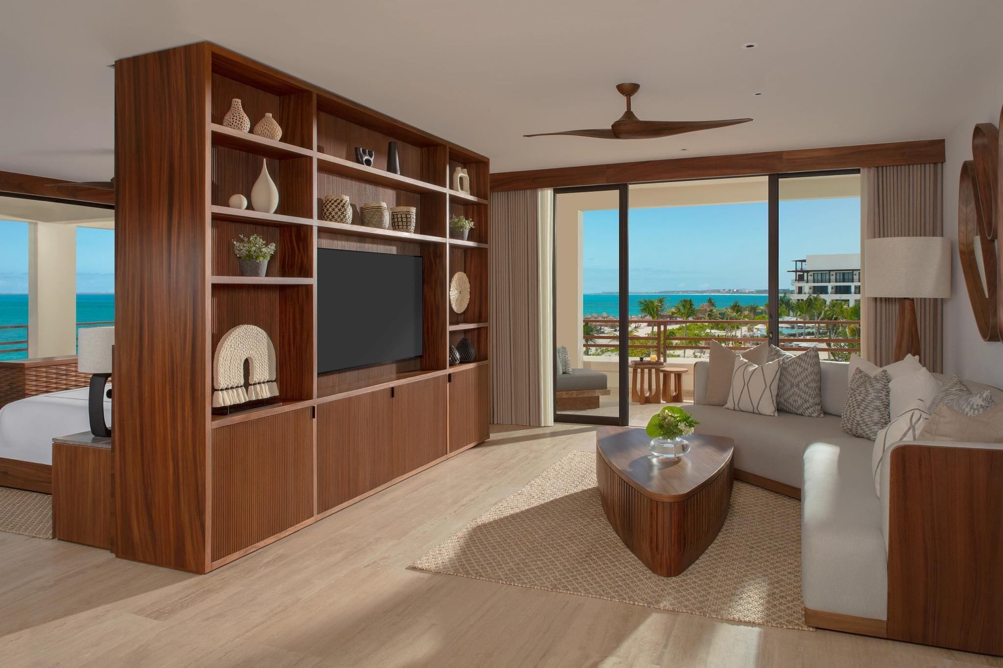 A modern living room with a large TV, wooden shelving, and a view of the ocean.