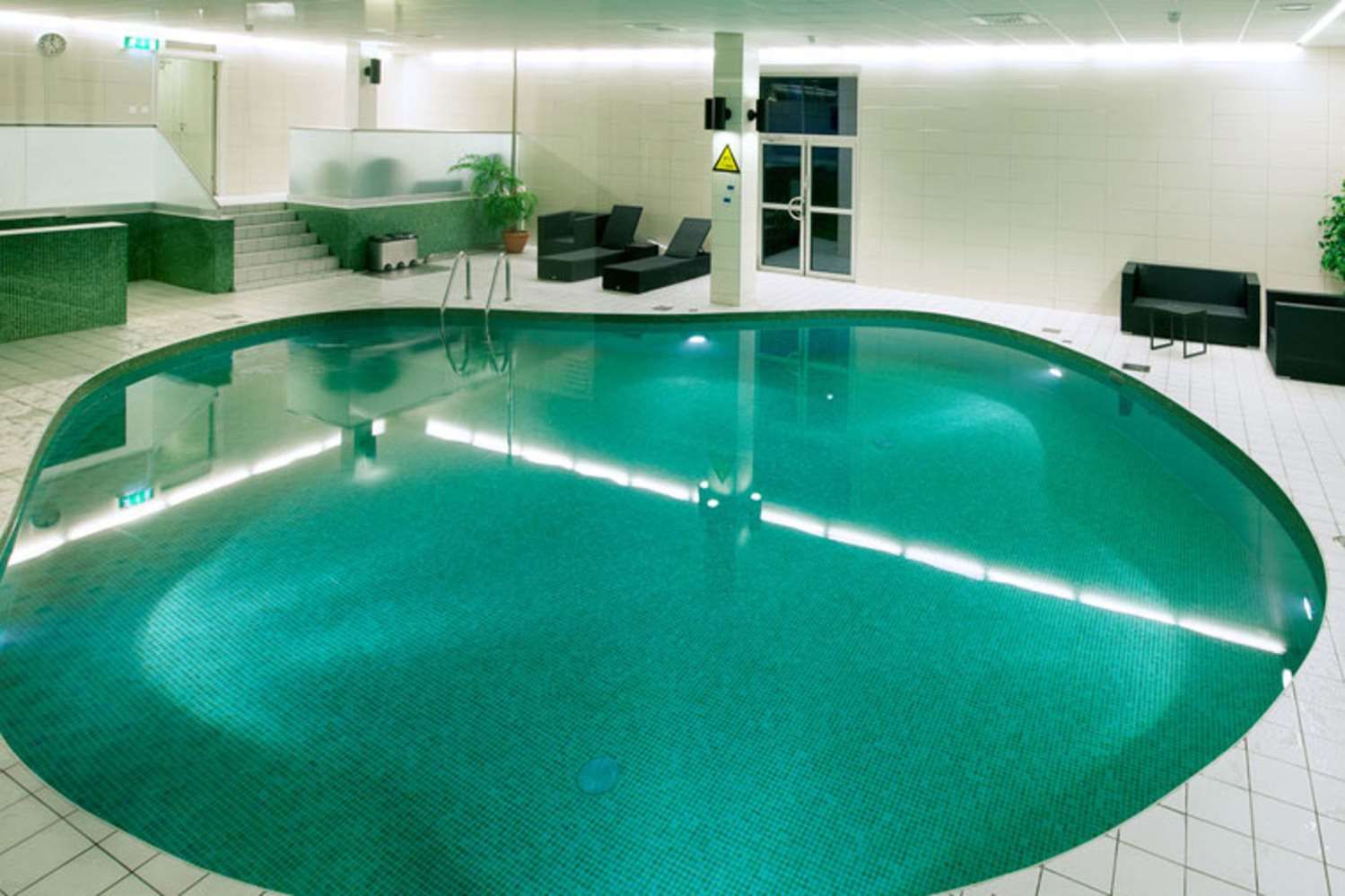 Una piscina interior con suelo de baldosas, una zona de estar con sillas y una mesa, y una escalera que lleva a un nivel superior.