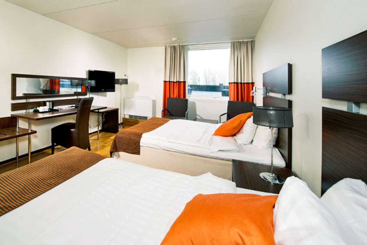 Habitación de hotel con cama, escritorio, silla, televisor y ventana con cortinas.