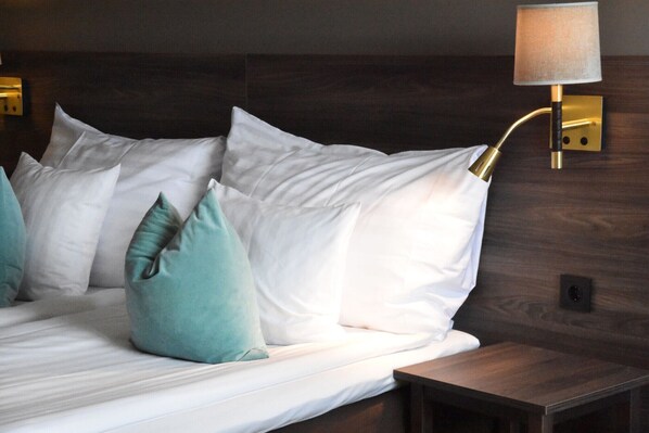 Una cama bien hecha con almohadas blancas y color turquesa, un cabecero de madera y una lámpara fijada a la pared.