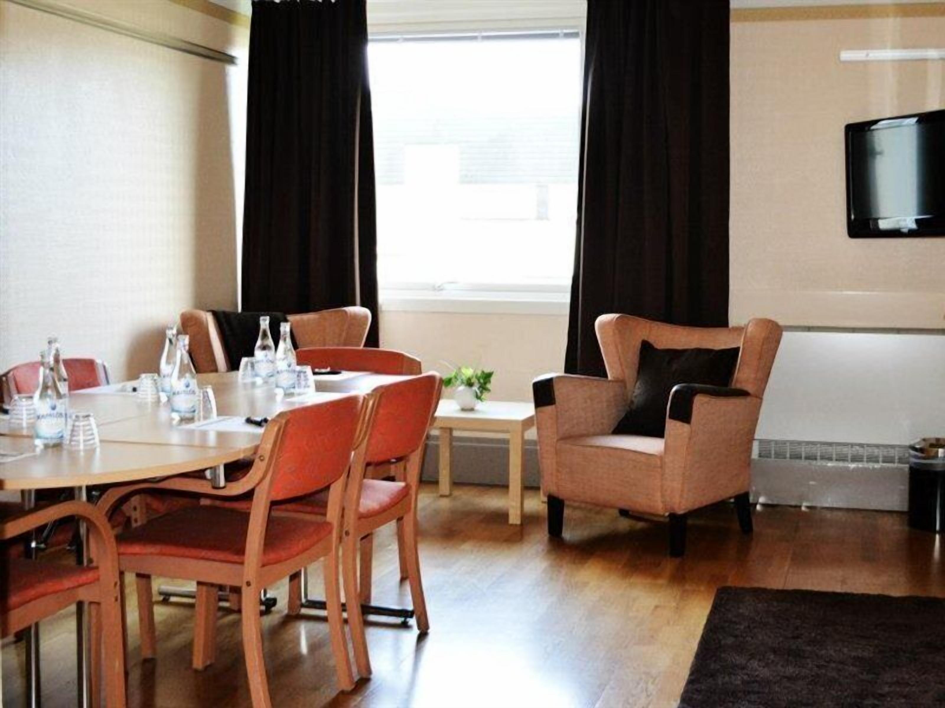 Habitación de hotel con una mesa de comedor, sillas, una pequeña mesita auxiliar, un sofá, un televisor montado en la pared y una ventana con cortinas.