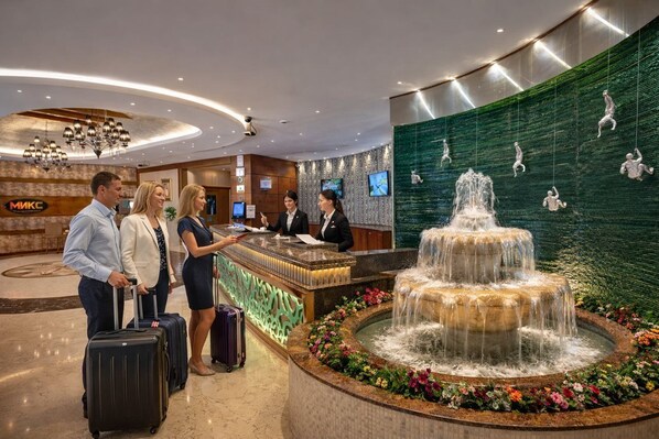 Zona de recepción de hotel con una fuente, mostrador de recepción y una pared con figuras trepando.