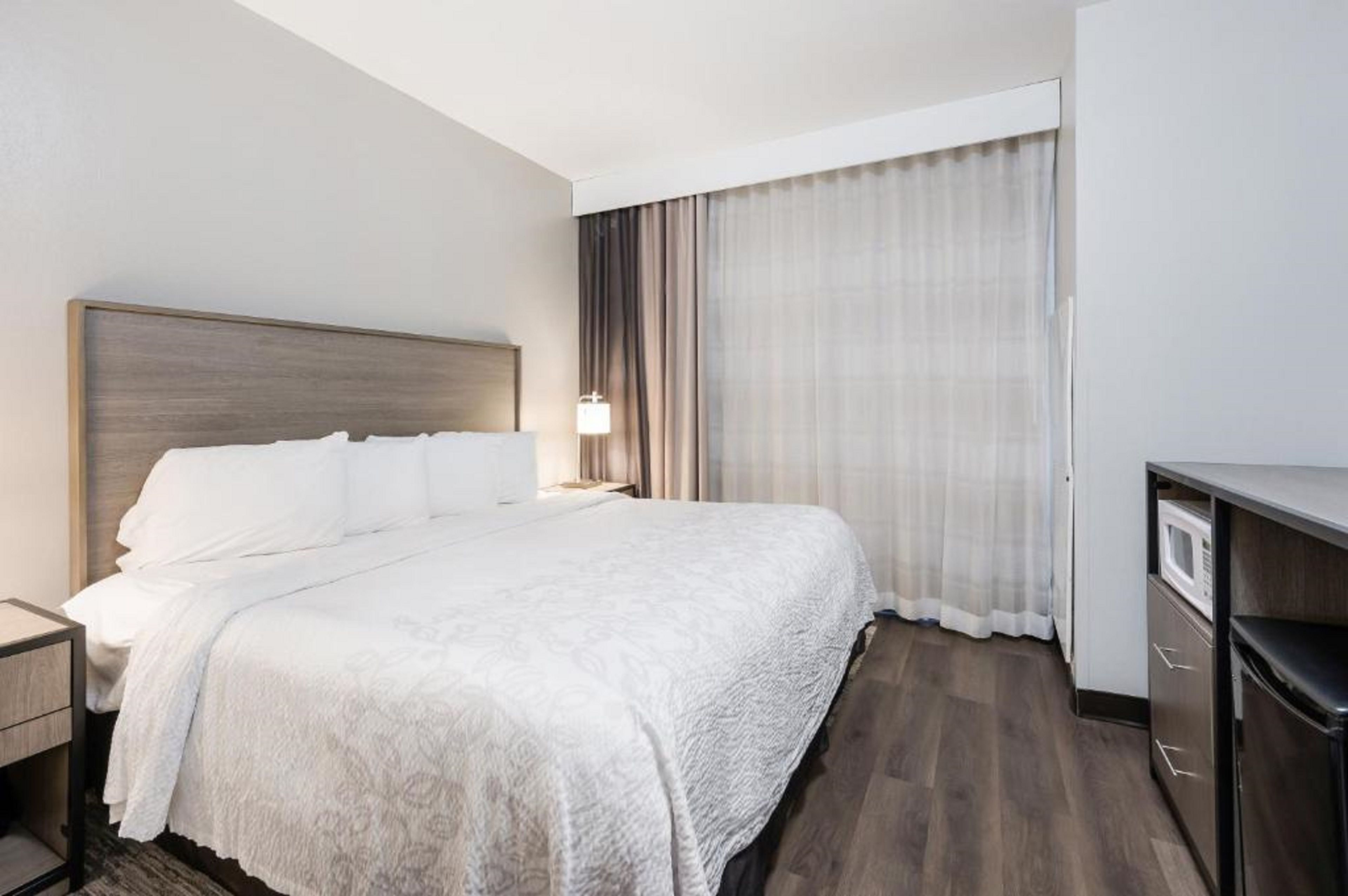 Habitación de hotel con cama, mesita de noche, lámpara, ventana con cortinas y microondas integrado.