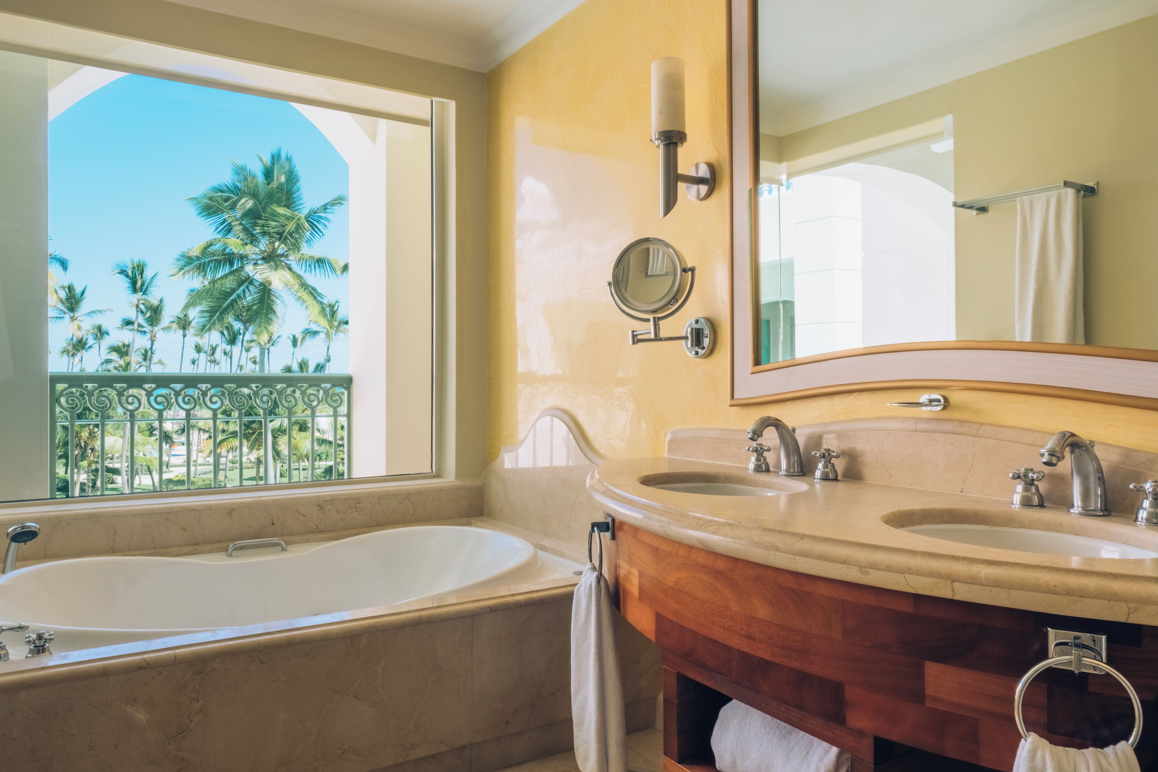 A bathroom with a bathtub, two sinks, a large mirror, and a view of palm trees through a window.