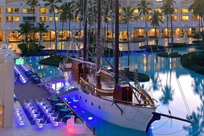 Una amplia piscina interior con una réplica de barco, rodeada de sillones y mesas, y un edificio de hotel al fondo.