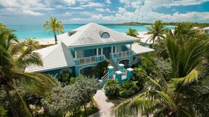Une maison turquoise à deux étages, dotée d’un balcon, entourée de palmiers et offrant une vue sur la plage.