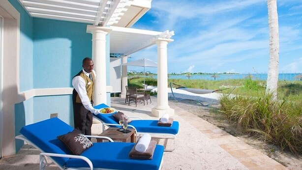 Un espace de détente aménagé sur la plage, avec des chaises longues bleues, un serveur apportant un repas et une vue sur l’océan.
