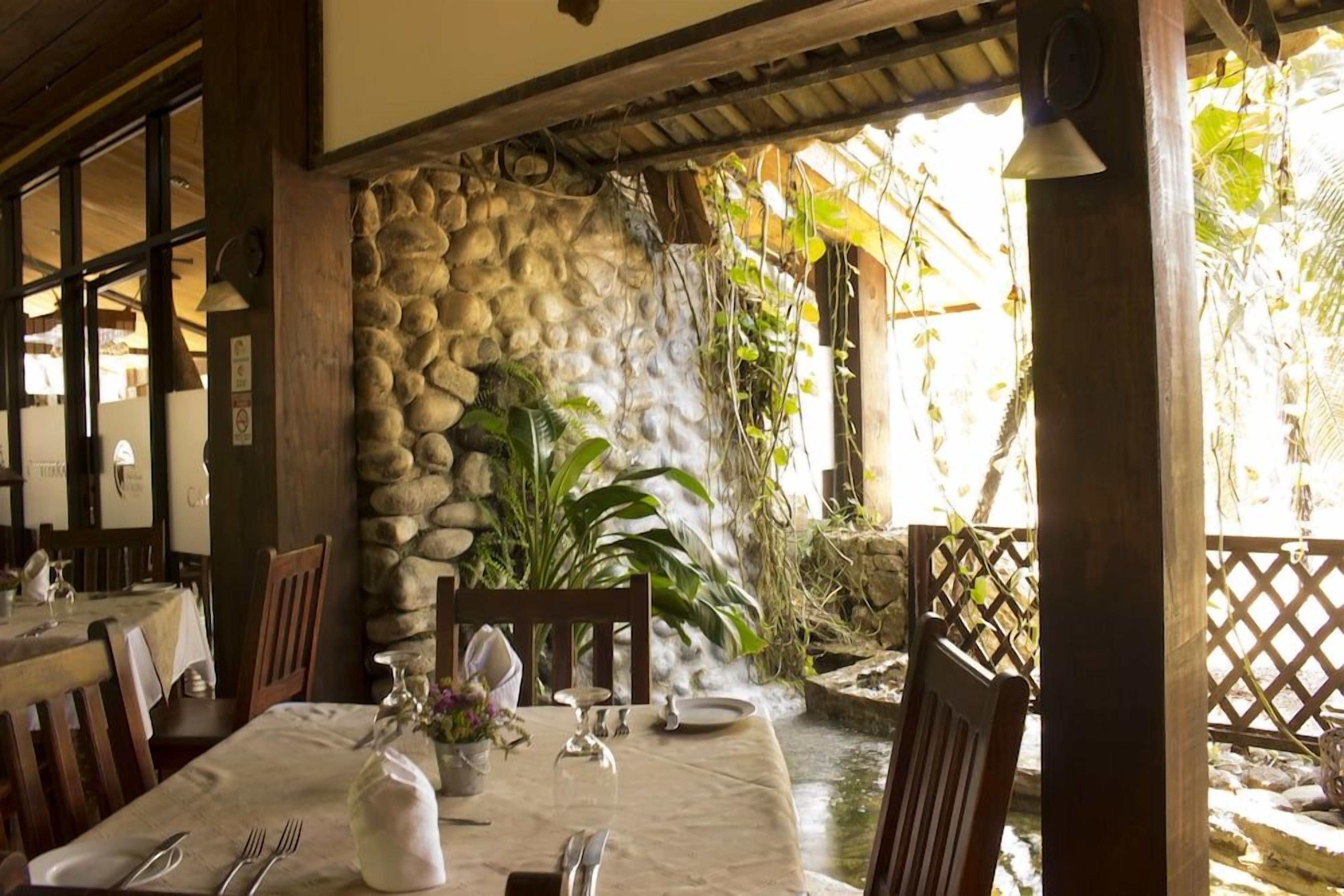 A restaurant with a stone wall, wooden furniture, and a view of a water feature.
