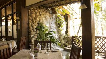 A restaurant with a stone wall, wooden furniture, and a view of a water feature.