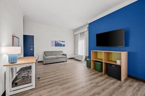 A modern living room with a sofa, a TV mounted on the wall, a small table with a lamp, and a kitchenette area. - Spark by Hilton Fort Myers (Fort Myers)