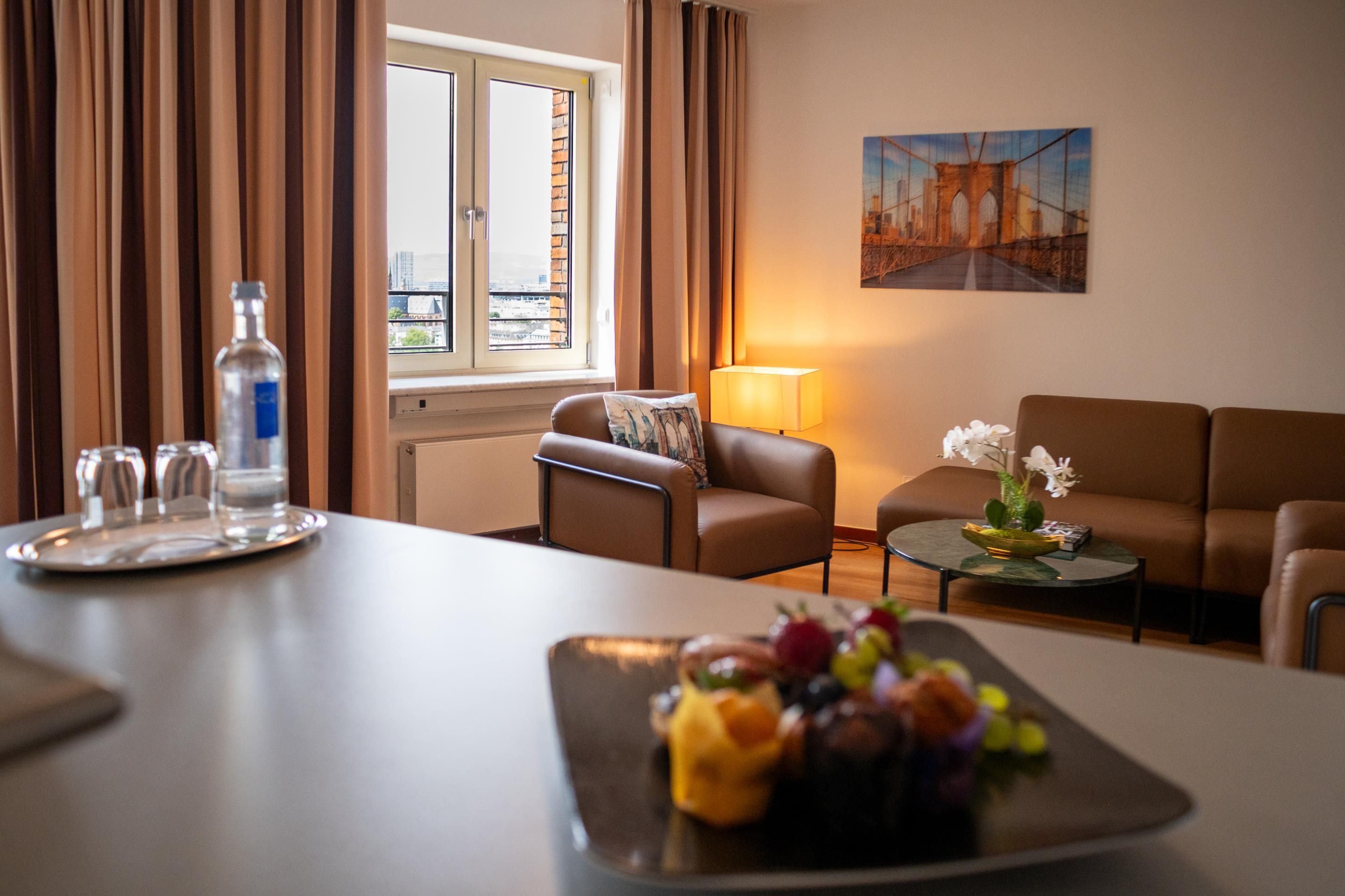 A hotel room with a dining table, a sofa, a small side table with a lamp, a painting on the wall, and a window with curtains.