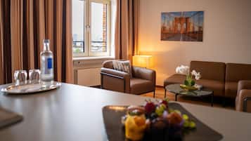 A hotel room with a dining table, a sofa, a small side table with a lamp, a painting on the wall, and a window with curtains.