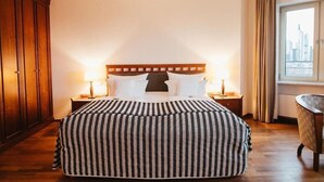 Une chambre d’hôtel avec un grand lit, une tête de lit en bois, deux tables de chevet avec des lampes, une chaise et une fenêtre avec des rideaux.