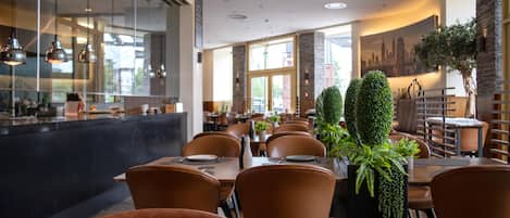 Interior de restaurante con mesas altas, sillas de cuero, iluminación colgante y un mural con vista a la ciudad.