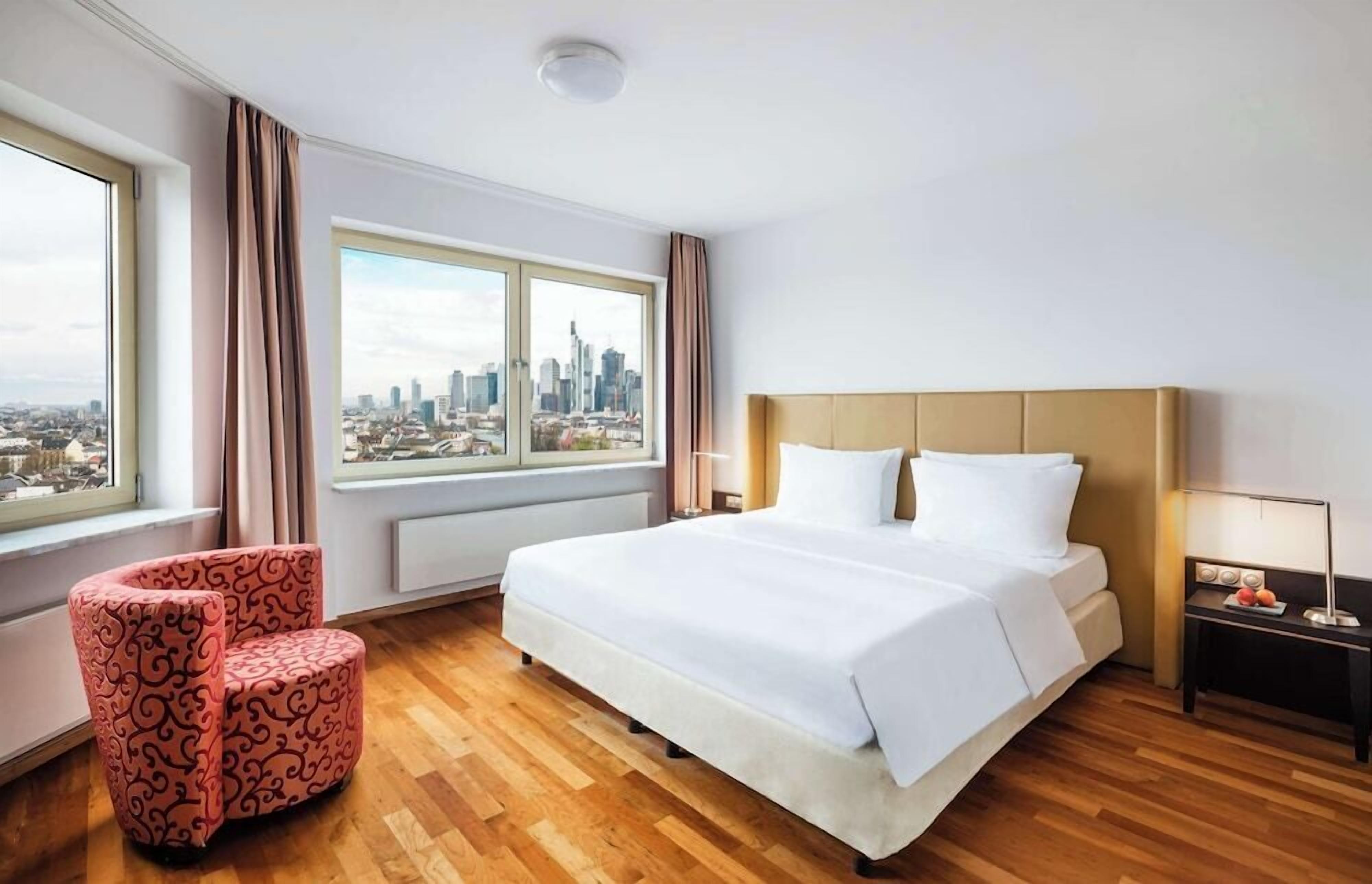 Ein Hotelzimmer mit einem großen Bett, einem Sessel, einem Nachttisch und einem Fenster mit Blick auf die Stadt.