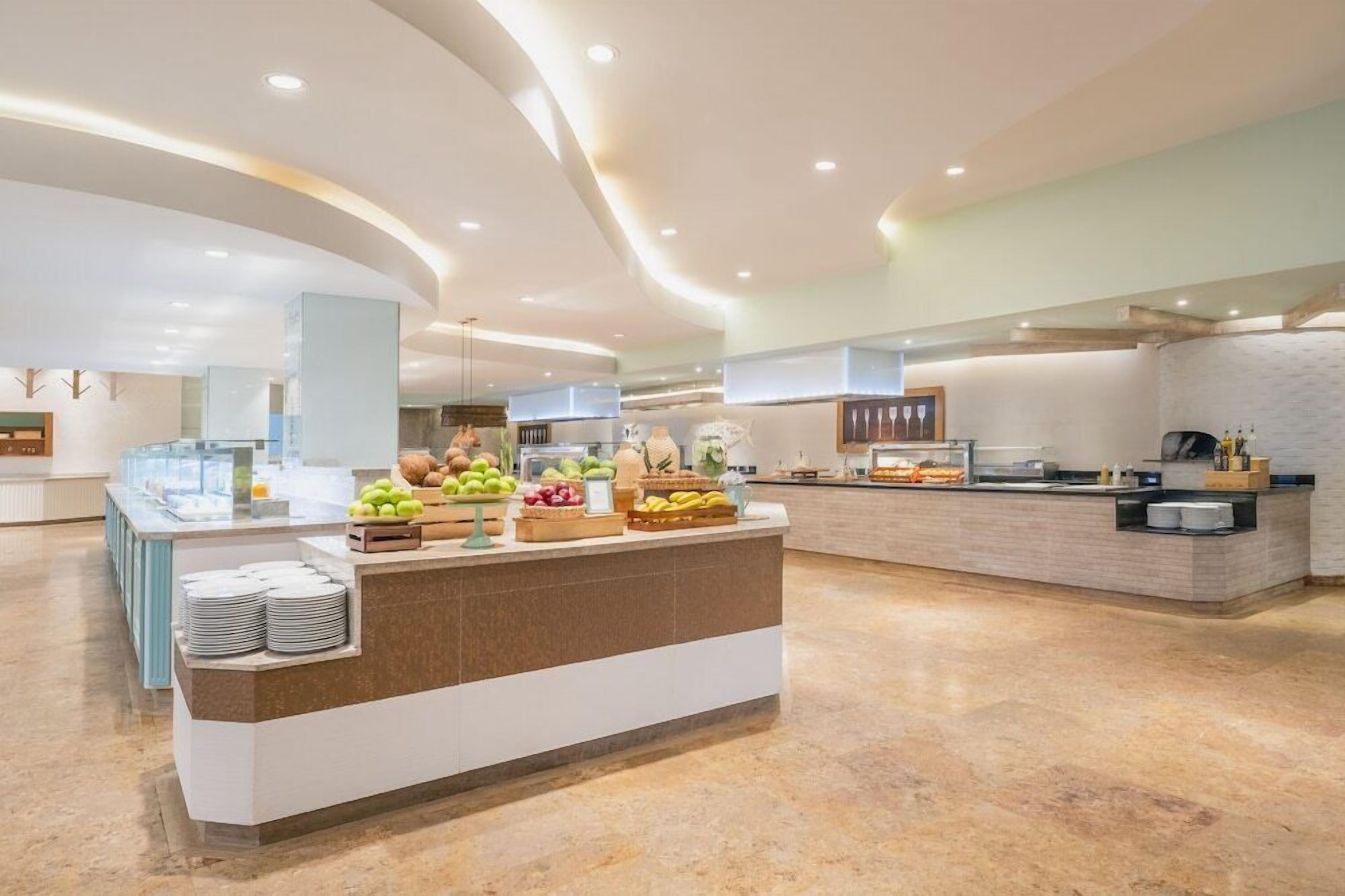 A modern hotel lobby with a central counter displaying fruits and beverages, a seating area with tables, and a decorative wall.