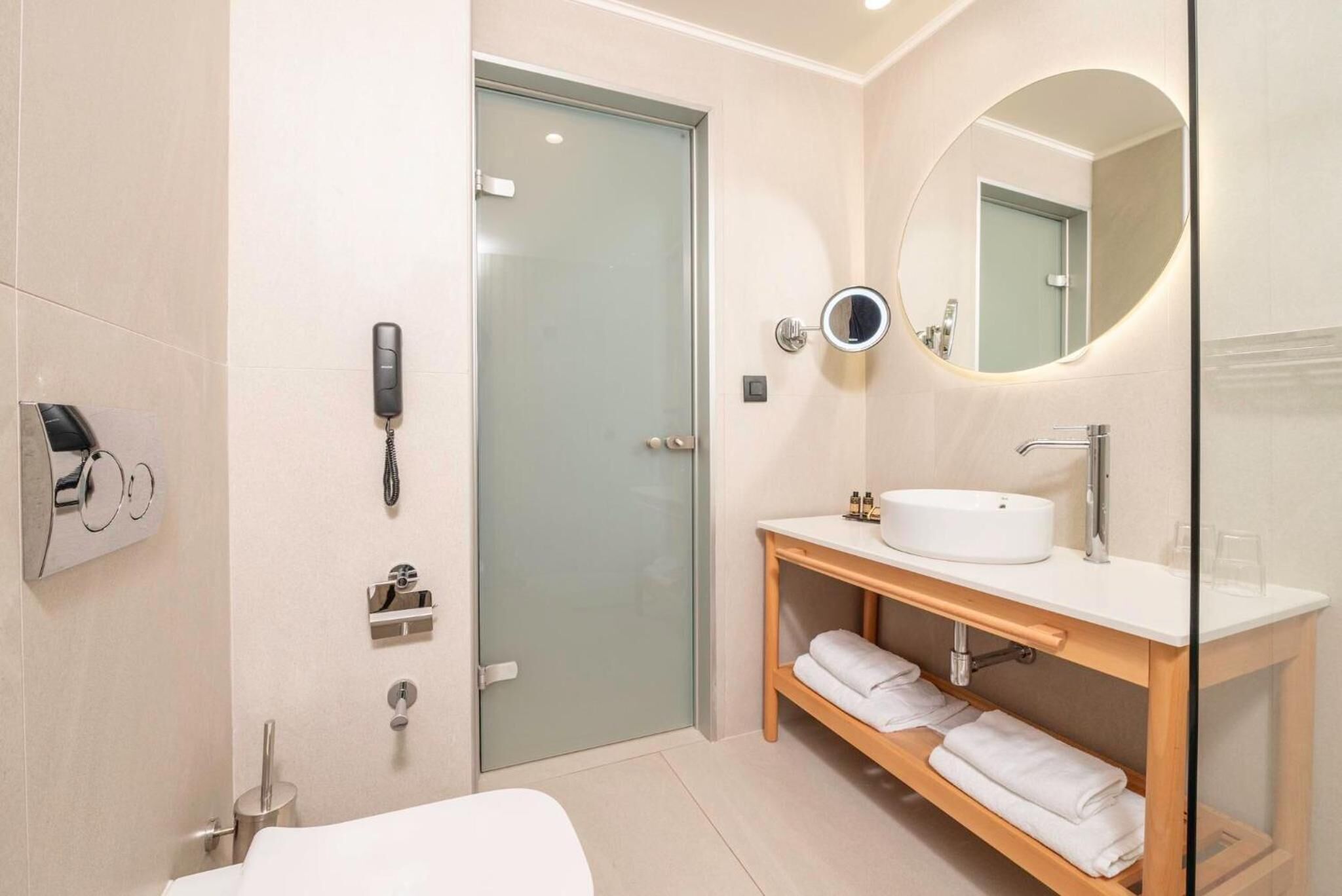Un baño moderno con cabina de ducha de vidrio, un lavamanos blanco, un espejo redondo y un mueble de tocador de madera con toallas.