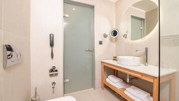 Un baño moderno con cabina de ducha de vidrio, un lavamanos blanco, un espejo redondo y un mueble de tocador de madera con toallas.