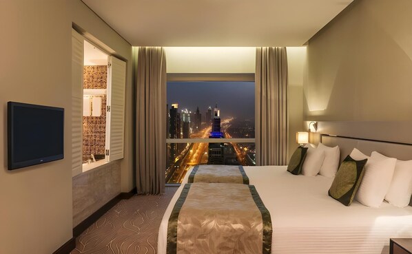 Habitación de hotel con cama, televisión, ventana con cortinas y vista a un paisaje urbano de noche.