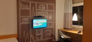 A hotel room with a TV mounted on a wall with a decorative design, a desk with a chair, a lamp, and a window with curtains. - Sure Hotel Spanga (Spånga)