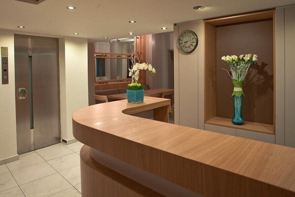 Un espace d’accueil d’hôtel avec un comptoir en bois incurvé, une horloge au mur et un vase de fleurs sur la droite.