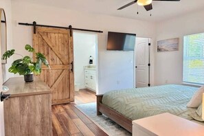 Une chambre à coucher comprenant un lit, une commode en bois, une porte coulissante de style rustique, un ventilateur de plafond et une fenêtre avec des stores.