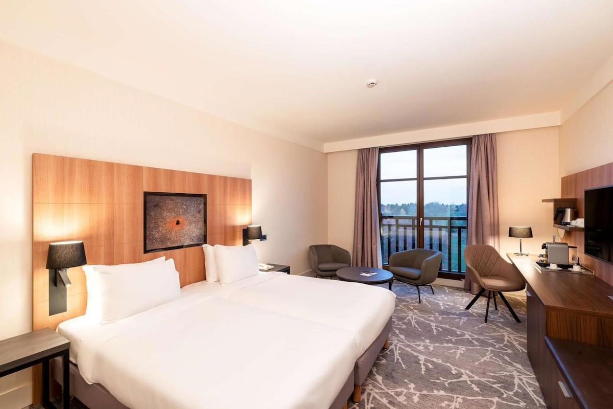 Een hotelkamer met een groot bed, een bureau met stoel, een televisie en uitzicht op het groen.