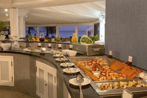 En buffet med forskellige spisemuligheder, herunder fisk og salater, i et godt oplyst rum med havudsigt.