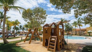 A wooden play structure with a ladder and swing set, surrounded by palm trees and a swimming pool. - TAN Resorts Marina Cap Cana (Punta Cana)