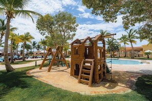 Eine Holzspielanlage mit Klettergerüst und Schaukeln, umgeben von Palmen und einem Swimmingpool.