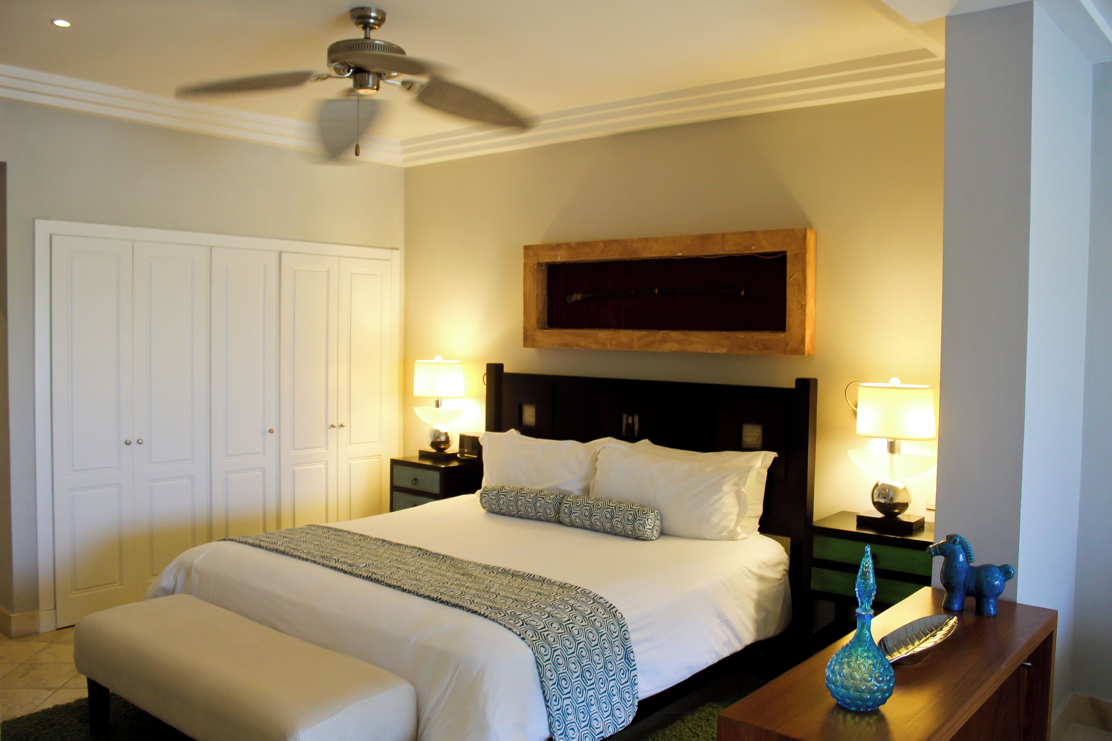Une chambre d’hôtel avec un grand lit, deux tables de chevet avec des lampes, un ventilateur de plafond et une tête de lit en bois.