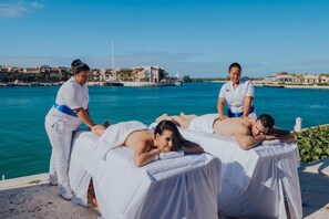 Ein Wellnessbereich mit Massagetischen, einer Wasserfläche und Gebäuden im Hintergrund.