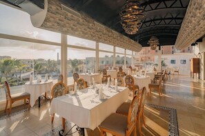A restaurant with multiple tables set for dining, large windows offering a view of the cityscape, and a high ceiling with decorative lighting. - TAN Resorts Marina Cap Cana (Punta Cana)