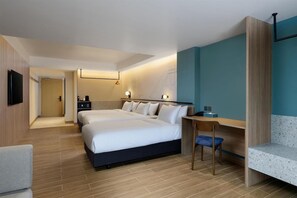 Habitación de hotel con una cama grande, un escritorio y una silla. Hay un televisor montado en la pared y una puerta que da a otra habitación.