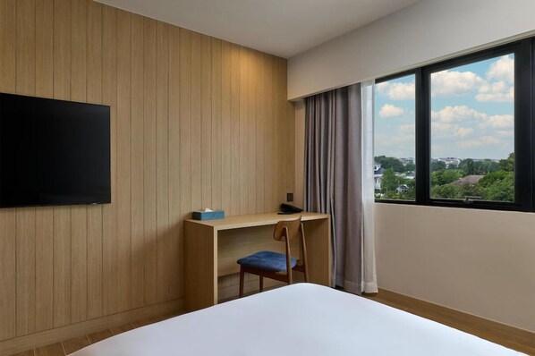 A hotel room with a bed, a desk with a chair, a flat-screen TV mounted on the wall, and a window with a view of the city. - Best Western Matter Tiwanon Hotel (Pak Kret)