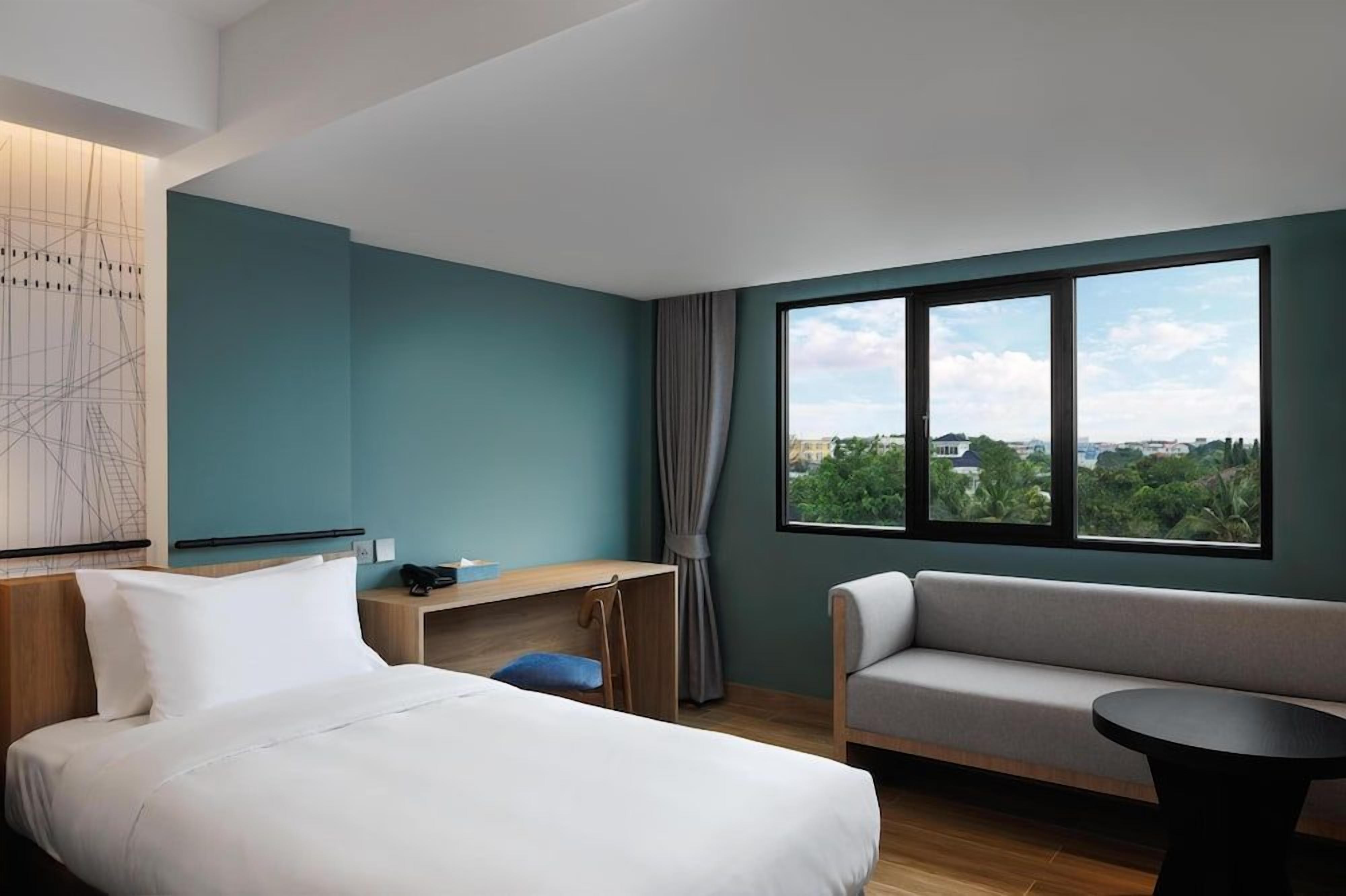 A hotel room with a bed, a desk, a sofa, and a window with a view of greenery and buildings.