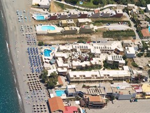 An aerial view of a coastal hotel with multiple swimming pools, beachside umbrellas, and a variety of building structures. - Indigo Mare (Platanias)